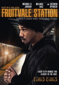 Image of a Black man standing in a subway station. He is wearing a black beanie and hoodie and has his hands clasped in front of him.
